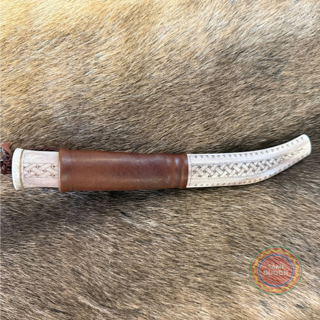 En sydsamisk halvhornskniv med sydsamisk gravyr av Same/Duodjislöjdare Sven-Åke Risfjells egen mönsterkomposition. Knivens hela längd i hölstret är 24 cm. Knivskaftet och hölstret är i renhorn. Det smidda bladets längd är 98 mm. Knivens upphängningsanordning är i tvinnat renskinn. ©Risfjells Sameslöjd i Vilhelmina södra Lappland.