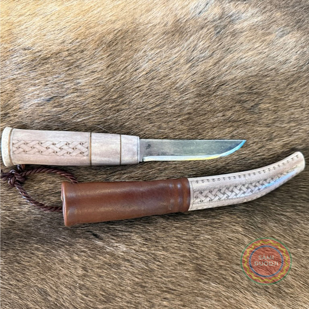 En sydsamisk halvhornskniv med sydsamisk gravyr av Same/Duodjislöjdare Sven-Åke Risfjells egen mönsterkomposition. Knivens hela längd i hölstret är 24 cm. Knivskaftet och hölstret är i renhorn. Det smidda bladets längd är 98 mm. Knivens upphängningsanordning är i tvinnat renskinn. ©Risfjells Sameslöjd i Vilhelmina södra Lappland.