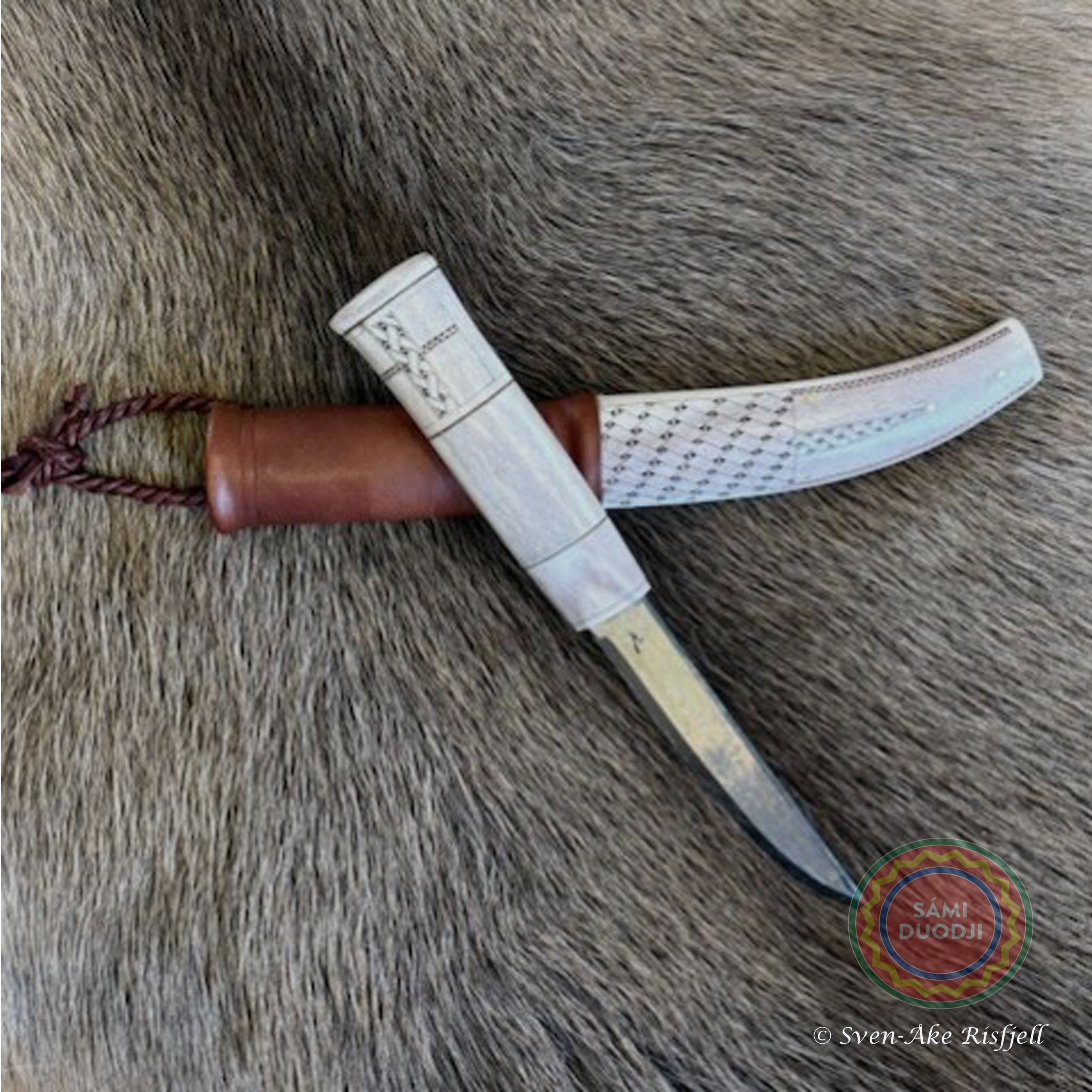 En sydsamisk halvhornskniv med sydsamisk gravyr av ©Same/Duodjislöjdare Sven-Åke Risfjells egen mönsterkomposition. Knivens hela längd i hölstret är 24 cm. Knivskaftet och hölstret är i renhorn. Det smidda bladets längd är 98 mm. Knivens upphängningsanordning är i tvinnat renskinn. Risfjells Sameslöjd i Vilhelmina södra Lappland.