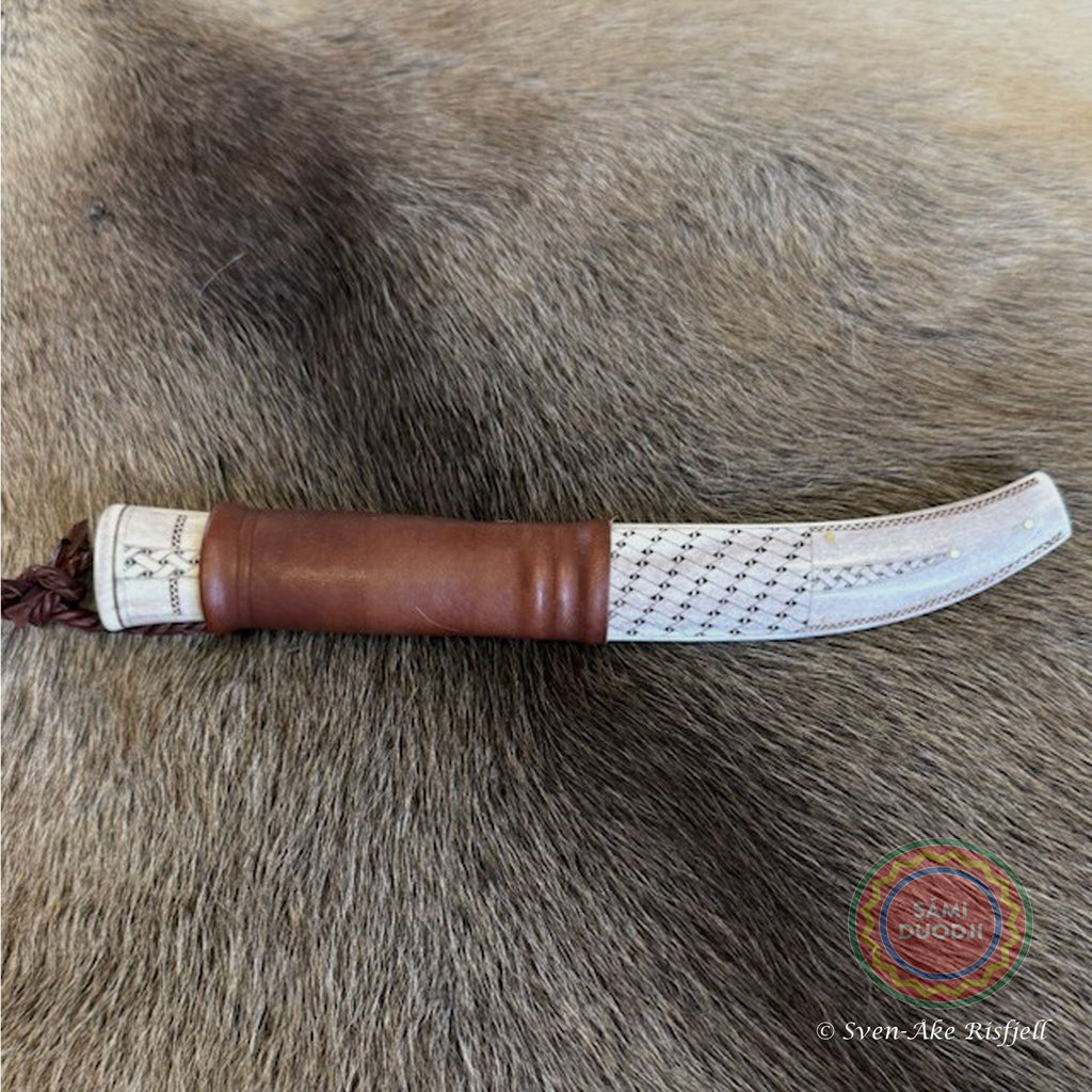 En sydsamisk halvhornskniv med sydsamisk gravyr av ©Same/Duodjislöjdare Sven-Åke Risfjells egen mönsterkomposition. Knivens hela längd i hölstret är 24 cm. Knivskaftet och hölstret är i renhorn. Det smidda bladets längd är 98 mm. Knivens upphängningsanordning är i tvinnat renskinn. Risfjells Sameslöjd i Vilhelmina södra Lappland.