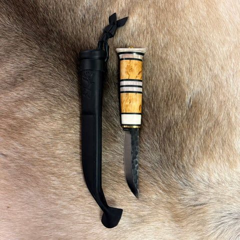 Jakt och fiskekniv (ej egen tillverkning) med hamrat blad blad 95 mm i kolstål. Knivskaftet gjord i masurbjörk och renhorn i fem fält. Nitad i topp. Mässing i nit och närmast knivbladet i skaftet. Svart läder i knivslidan som har en ren tryckt som dekoration. Knivens längd 21 cm och med hölster 25 cm. Hos Risfjells Sameslöjd