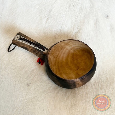 En kåsa ur björkvril med horninlägg i skaftet av same/Duodjislöjdare Sven-Åke Risfjell. Gravyren är av sydsamisk tradition. Kåsan rymmer 1,5-2 dl. Koppens diameter 9 cm. Kåsans längd är 15,5 cm och höjd 5 cm. Tvinnad rem av renskinn med tofs i kläde i rött och grönt. Risfjells Sameslöjd i Vilhelmina, södra Lappland