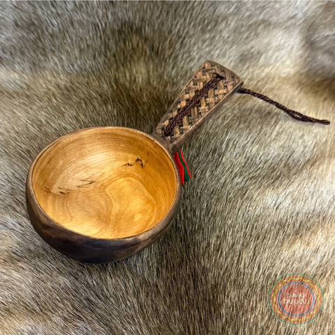 Kåsa ur björkvril med karvsnittsgravyr med sydsamiskt mönster på skaftets ovansida&nbsp; och undersidan av skålen på kåsan. Tillverkad av same/Duodjislöjdare Sven-Åke Risfjell. Kåsan rymmer lite drygt 1,5 dl. Koppens diameter 9 cm. Kåsans längd är 16 cm. Djup 4,5 cm. Tvinnad rem av renskinn med tofs i kläde i rött och grönt. Risfjells Sameslöjd i Vilhelmina, södra Lappland