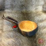 Kåsa ur björkvril med riklig karvsnittsgravyr med sydsamiskt mönster på skaftets under och ovansida&nbsp; och undersidan av skålen på kåsan. Tillverkad av same/Duodjislöjdare Sven-Åke Risfjell. Kåsan rymmer lite drygt 1,5 dl. Koppens diameter 8,5 cm. Kåsans längd är 15,5 cm. Djup 3,5 cm. Tvinnad rem av renskinn med tofs i kläde i rött och grönt. Risfjells Sameslöjd i Vilhelmina, södra Lappland