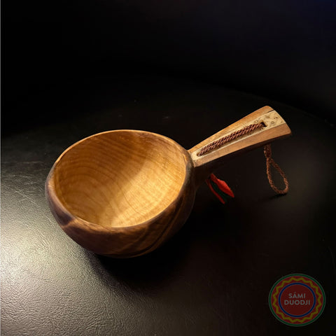 En kåsa ur björkvril med horninlägg i skaftet av same/Duodjislöjdare Sven-Åke Risfjell. Gravyren är av sydsamisk tradition. Kåsan rymmer 1,5-2 dl. Koppens diameter 9 cm. Kåsans längd är 15,6 cm och höjd 4,5 cm. Tvinnad rem av renskinn med tofs i kläde i rött och grönt. Risfjells Sameslöjd i Vilhelmina, södra Lappland