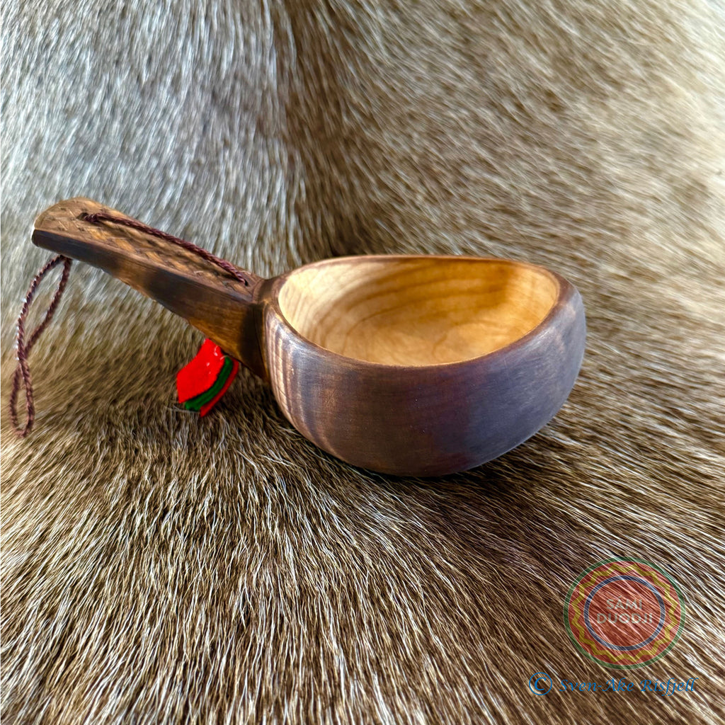 Kåsa ur björkvril med riklig karvsnittsgravyr med sydsamiskt mönster på skaftets under och ovansida&nbsp; och undersidan av skålen på kåsan. Tillverkad av same/Duodjislöjdare Sven-Åke Risfjell. Kåsan rymmer lite drygt 1,5 dl. Koppens diameter 9,5 cm. Kåsans längd är 16,5 cm. Höjd 5 cm. Tvinnad rem av renskinn med tofs i kläde i rött och grönt. Risfjells Sameslöjd i Vilhelmina, södra Lappland