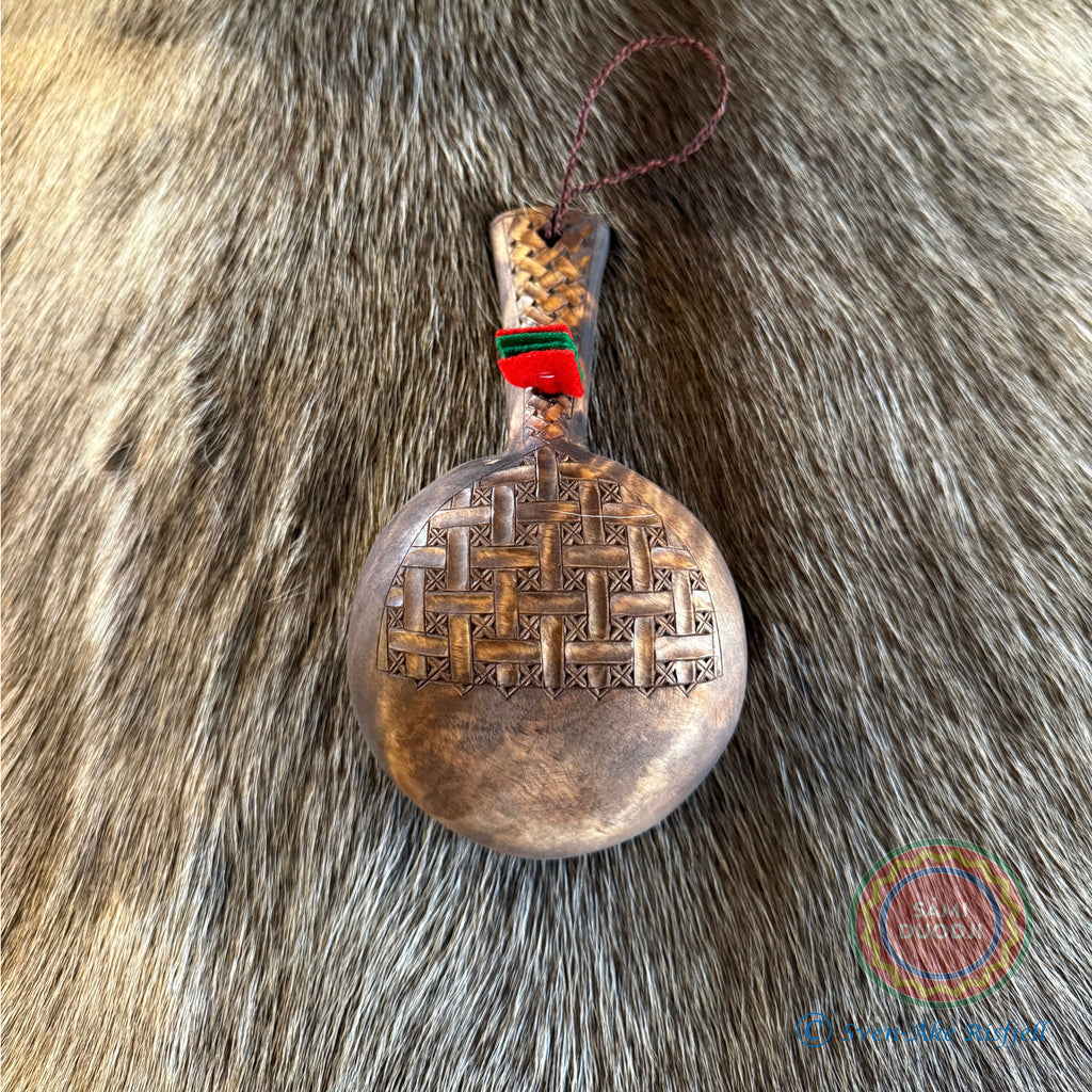 Kåsa ur björkvril med riklig karvsnittsgravyr med sydsamiskt mönster på skaftets under och ovansida&nbsp; och undersidan av skålen på kåsan. Tillverkad av same/Duodjislöjdare Sven-Åke Risfjell. Kåsan rymmer lite drygt 1,5 dl. Koppens diameter 9,5 cm. Kåsans längd är 16,5 cm. Höjd 5 cm. Tvinnad rem av renskinn med tofs i kläde i rött och grönt. Risfjells Sameslöjd i Vilhelmina, södra Lappland