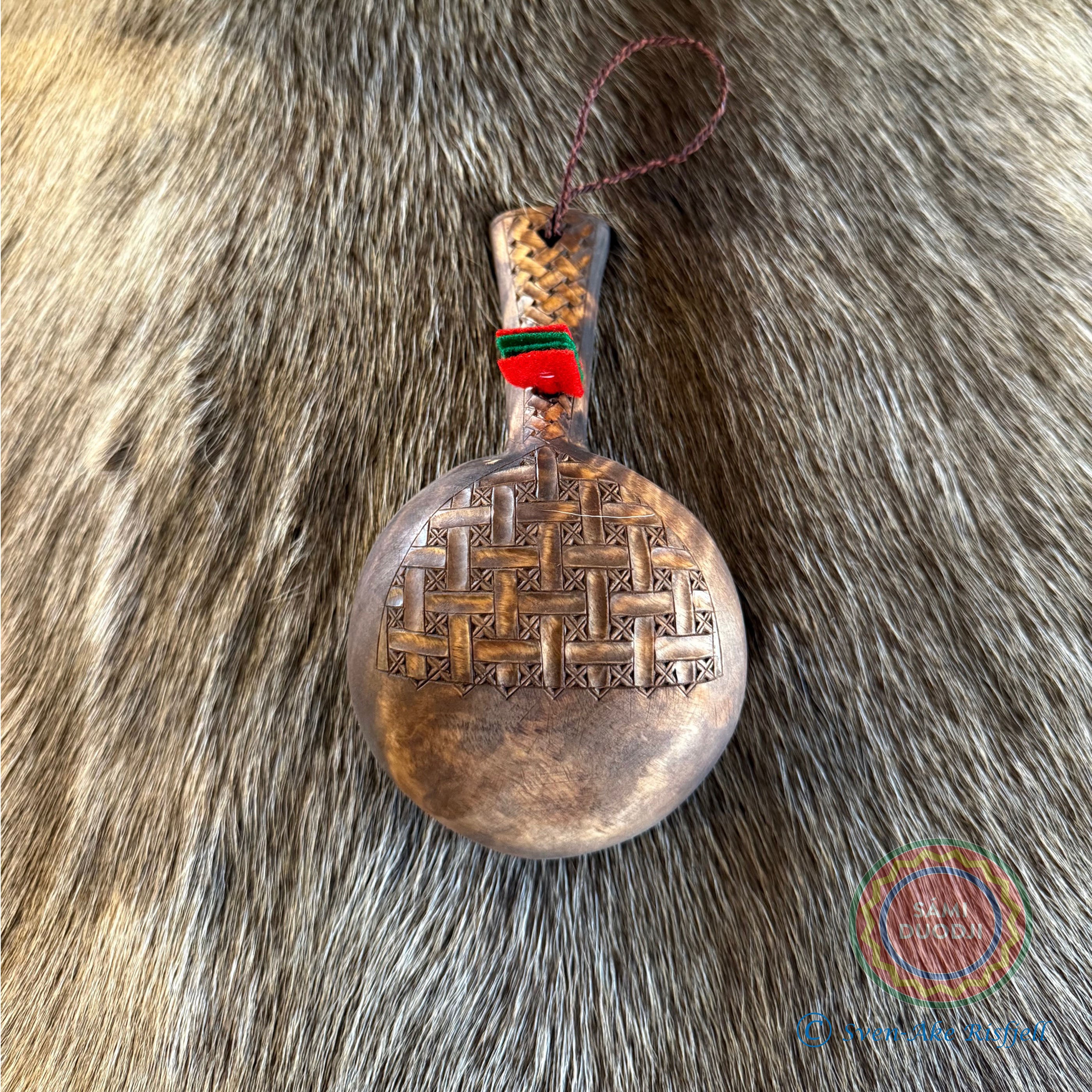 Kåsa ur björkvril med riklig karvsnittsgravyr med sydsamiskt mönster på skaftets under och ovansida&nbsp; och undersidan av skålen på kåsan. Tillverkad av same/Duodjislöjdare Sven-Åke Risfjell. Kåsan rymmer lite drygt 1,5 dl. Koppens diameter 9,5 cm. Kåsans längd är 16,5 cm. Höjd 5 cm. Tvinnad rem av renskinn med tofs i kläde i rött och grönt. Risfjells Sameslöjd i Vilhelmina, södra Lappland