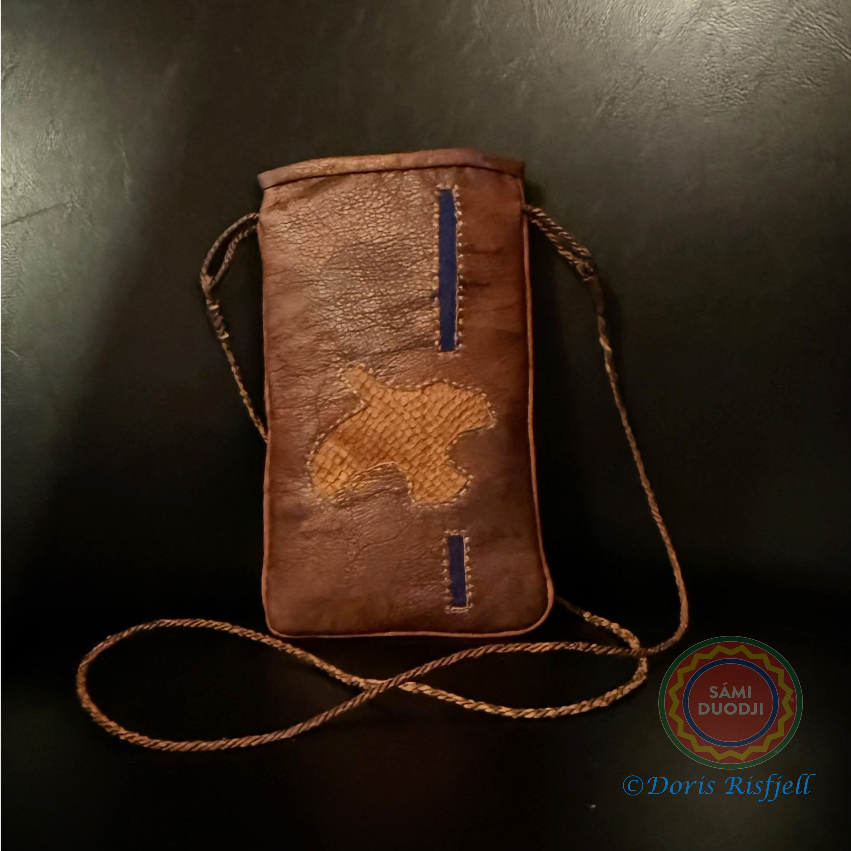 Mobilväska eller varför inte en liten väska i renskinn när du bara behöver mobil och betalkort med dig. Av sameslöjdare Doris Risfjell Vilhelmina. Väskan är dekorerad med en flygande ripa i brunt laxskinn och inlägg av blått kläde. Höjd 17cm och bredd 10 cm. Väskan har blått foder och en innerficka. Stängningsanordning med renhornsknapp och tvinnad renskinnsögla. Tvinnad rem i renskinn som mäter 45 cm från väskans övre kant till halsen. Risfjells Sameslöjd Vilhelmina.