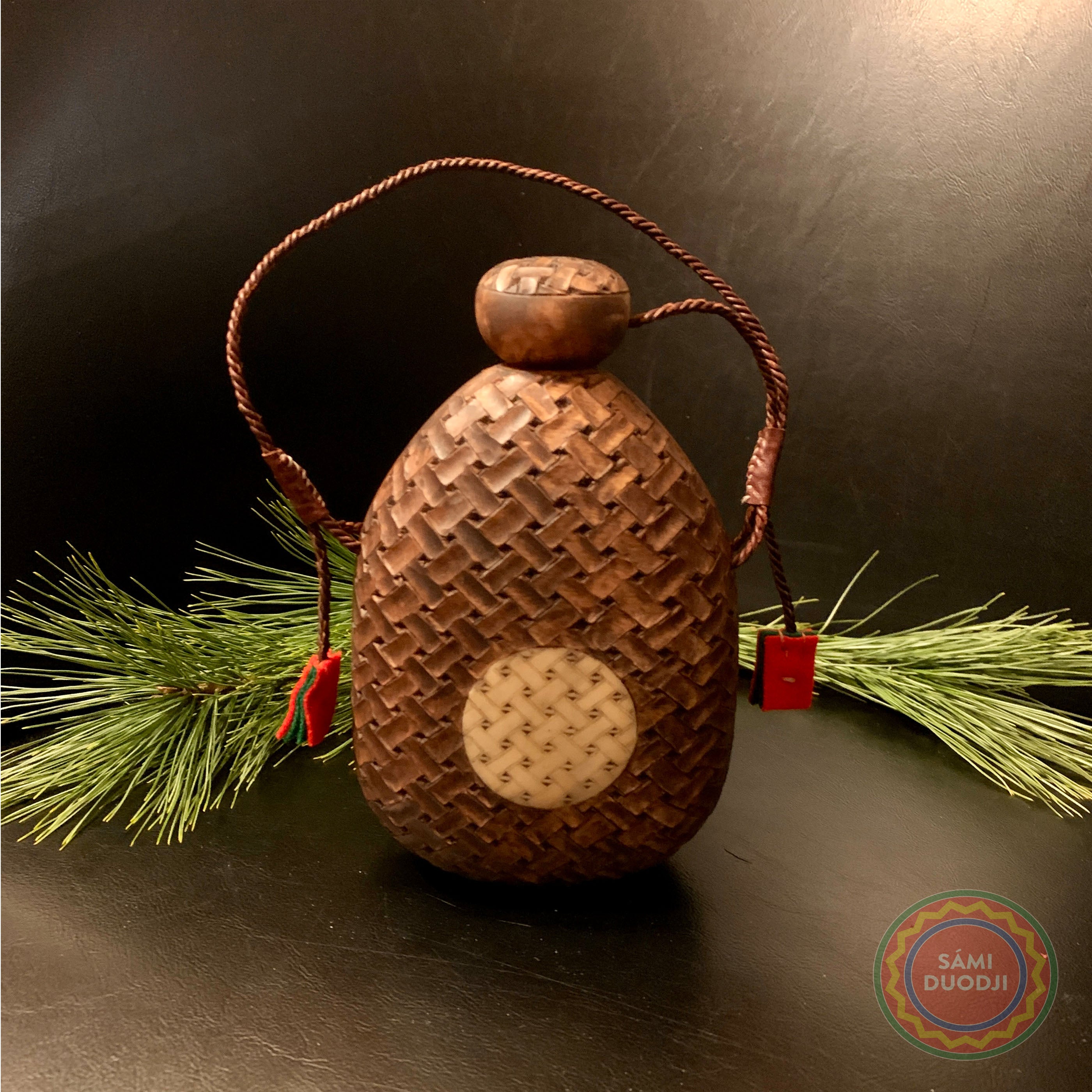 Samisk saltflaska ur björkvril med karvsnittsgravyr av same-Duodjislöjdare Sven-Åke Risfjell Vilhelmina. Flaskan har horninlägg med sydsamisk gravyr på framsidan. Karvsnittsgravyren tog Sven-Åke upp på allvar från det han firade sitt 25 års jubileum som Duodjislöjdare. Flaskans höjd är 17 cm och bredden är 11,5 cm. Tvinnad läderrem som handtag där små klädestofsar i rött och grönt ger det lilla extra i utförandet. Risfjells Sameslöjd i Vilhelmina Södra Lappland