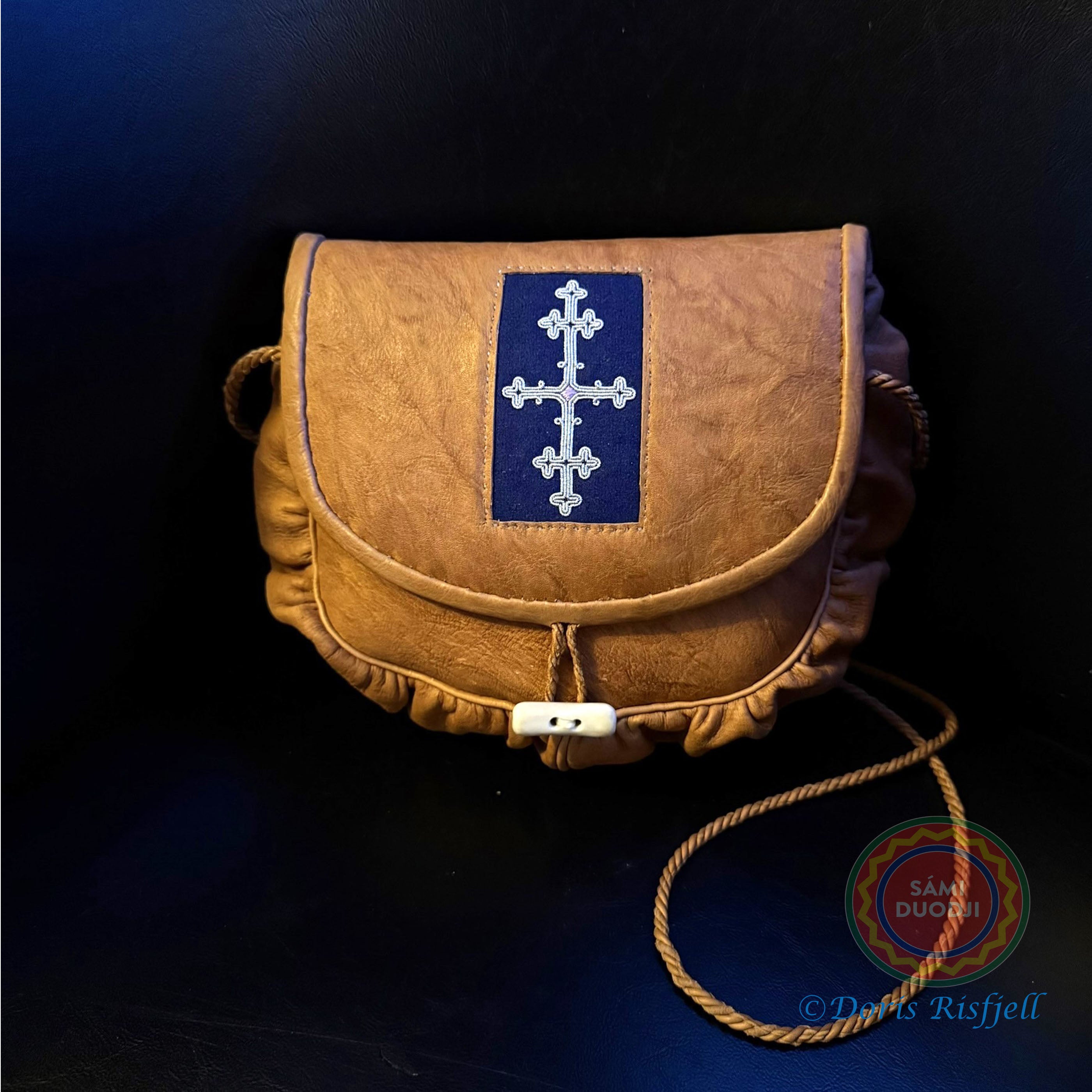 Väskan är sydd i hemgarvat hemgarvat renskinn i traditionell samisk design av same/duodjislöjdare Doris Risfjell. Väskan är dekorerad med tenntrådsbrodyr på blått kläde. Den är 22 cm bred och 18 cm hög. Axelremmen som är av tvinnat renskinn mäter 62 cm från väskans övre kant till axeln. Väskans insida är fodrad i blått och har en innerficka. Renhornsknapp med tvinnad renskinnsögla i stängningsanordningen. Risfjells Sameslöjd i Vilhelmina.