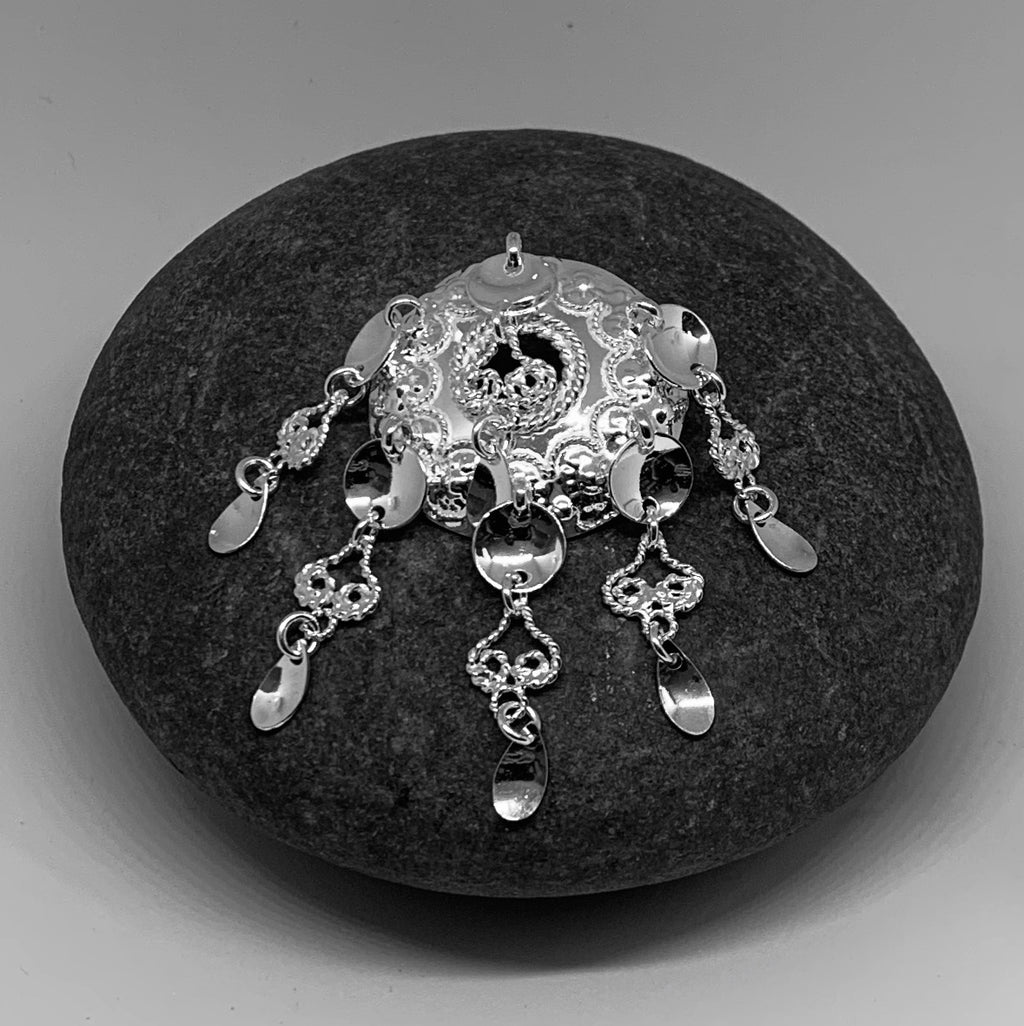 Samiskt silverbrosch med rik utsmyckning. Runda och skedformade löv på sex hängen. Diameter på bucklan som är punsad och dekorerad med filigranarbete är 30 mm. Hängena är 32 mm långa.  Broschet är en kopia på Sven-Åke Risfjells farmor Sara Alvina Sofia Sjulsdotters brosch. Broschet är fotat på Sara Avinas kaffesten med vilken hon malde sina kaffebönor. finns hos Risjfells Sameslöjd i Vilhelmina.