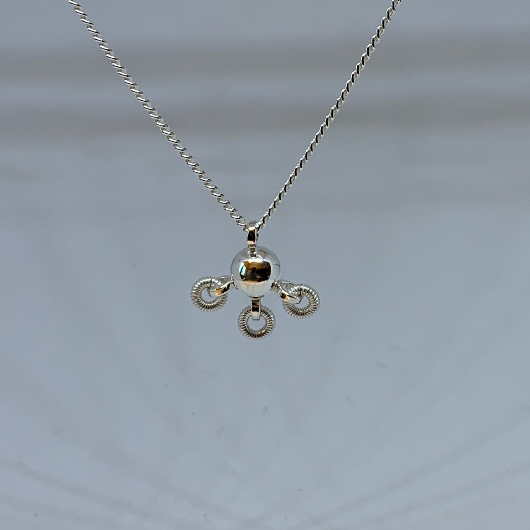 Söt komsekula i silver med tre små lindade ringar hängande på undersidan av kulan. Silverkedjan är 42 cm lång. Kulans diameter är 7 mm. Komsekulan är ett samiskt kultursmycke. Finns hos Risfjells Sameslöjd i Vilhelmina, södra Lappland.