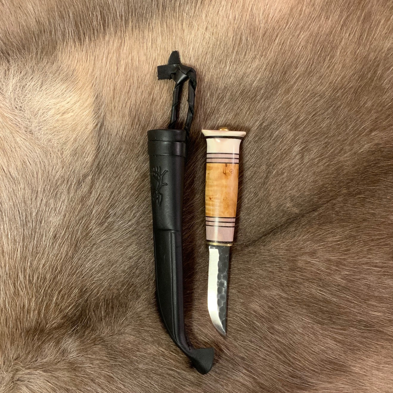 Jakt och fiskekniv (ej egen tillverkning)med hamrat blad 95 mm i kolstål. Knivskaftet gjord i masurbjörk och renhorn, nitad i topp. Mässing i nit och närmast knivbladet i skaftet. Skaftet har två fält horn med sex mörka inlägg.Svart läder i knivslidan som har en ren tryckt som dekoration. Knivens längd 21 cm och med hölster 25 cm. Finns hos Risjfells Sameslöjd i Vilhelmina