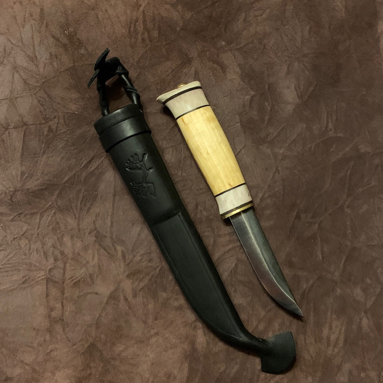  Jakt och fiskekniv (ej egen tillverkning)med slätt blad 95 mm i kolstål. Knivskaftet gjord i masurbjörk och renhorn i två fält. Nitad i topp. Mässing i nit och närmast knivbladet i skaftet. Svart läder i knivslidan som har en ren tryckt som dekoration. Knivens längd 21 cm och med hölster 25 cm. Finns hos Risfjells Sameslöjd i Vilhelmina.