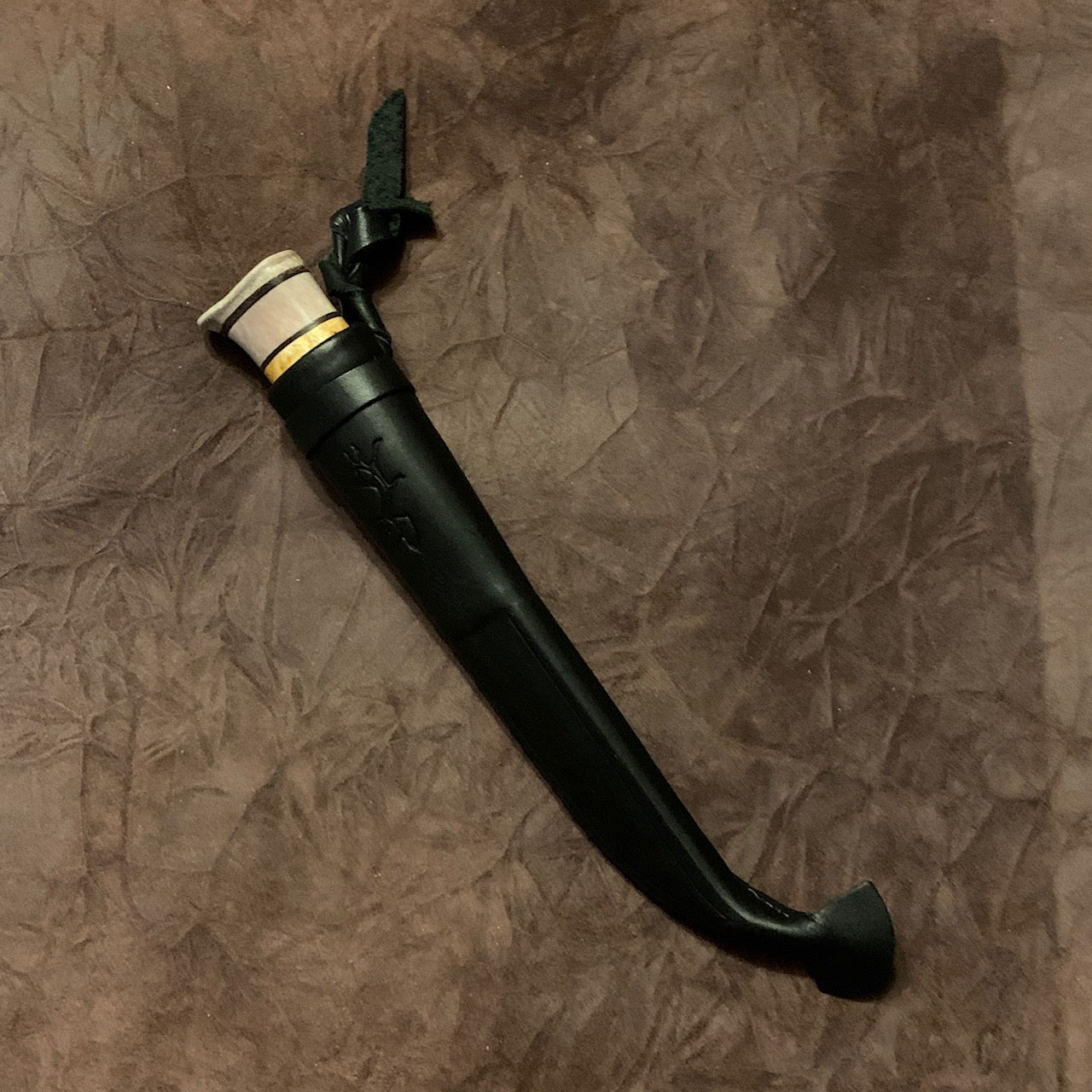 Kniv i masurbjörk och tre fält horn med nitad horntopp och svart läderslida. Knivens hela längd i hölster mäter 25 cm. Knivens blad har en dekorativ hamrad översida och är är 90 mm. Ej egen tillverkning. Bra och vacker allt i allo kniv. Finns hos Risjfells Sameslöjd i Vilhelmina.