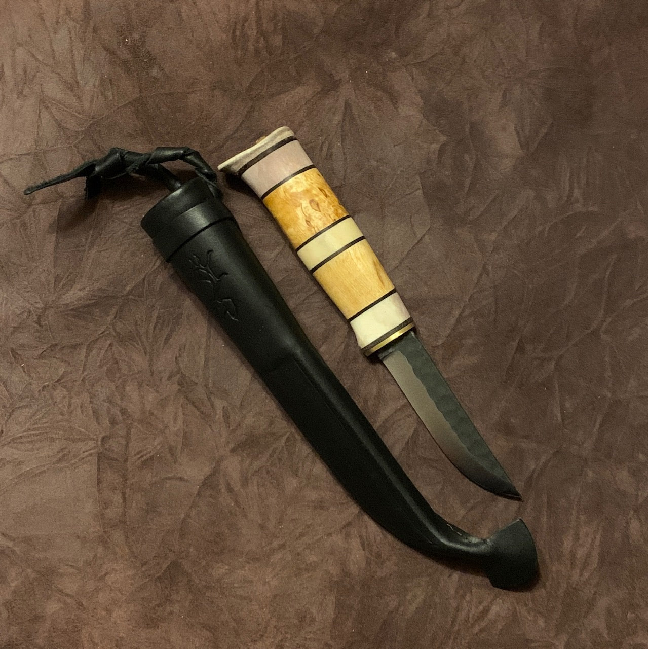 Kniv i masurbjörk och tre fält horn med nitad horntopp och svart läderslida. Knivens hela längd i hölster mäter 25 cm. Knivens blad har en dekorativ hamrad översida och är är 90 mm. Ej egen tillverkning. Bra och vacker allt i allo kniv. Finns hos Risjfells Sameslöjd i Vilhelmina.