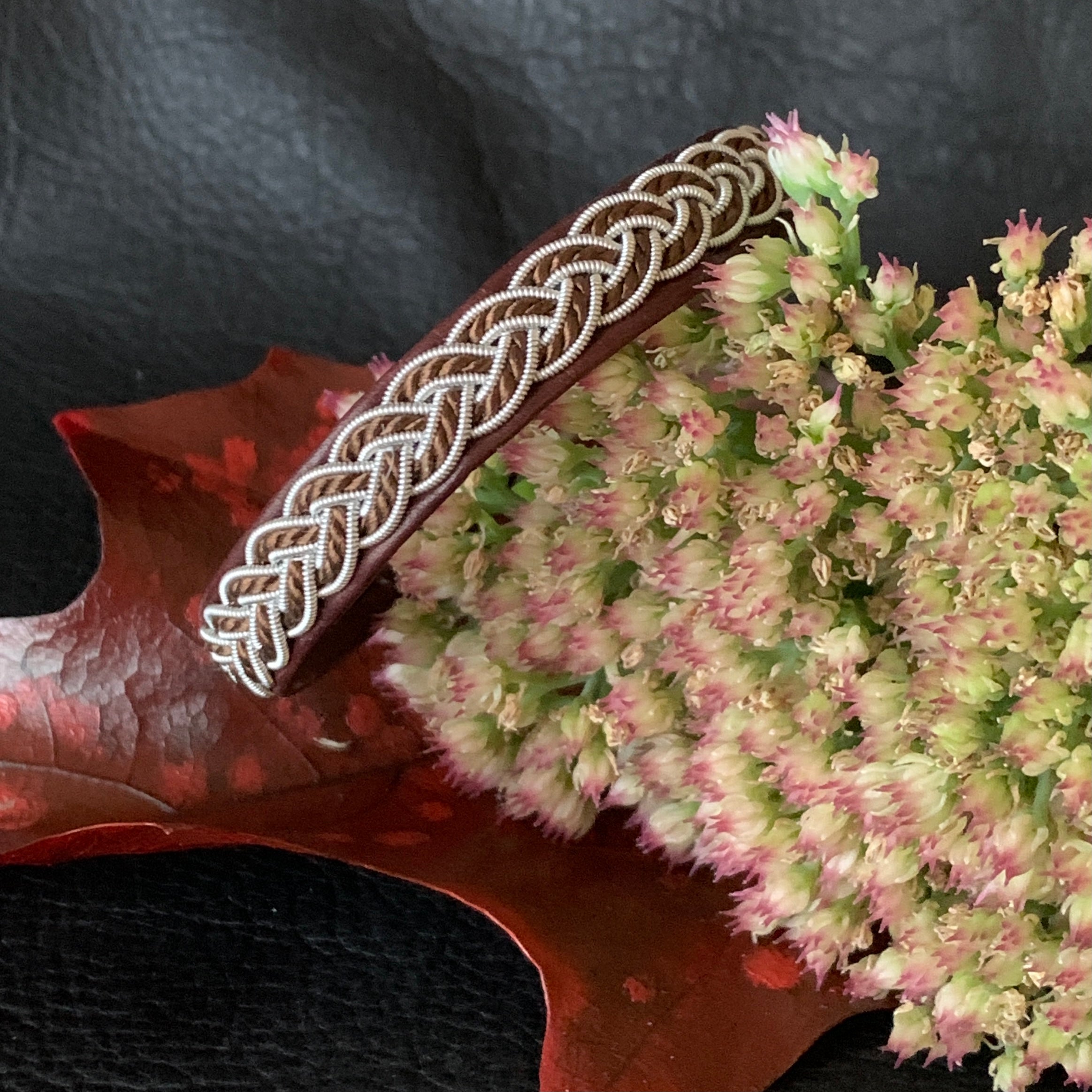 Tenntrådsarmband med flätning av tenntråd och tvinnat mörkbrunt garn på brunt renskinn. Knapp i renhorn och ögla i tvinnat renskinn. Bredd ca 10 mm. Samiskt hantverk av Doris Risfjell. Risfjells Sameslöjd.