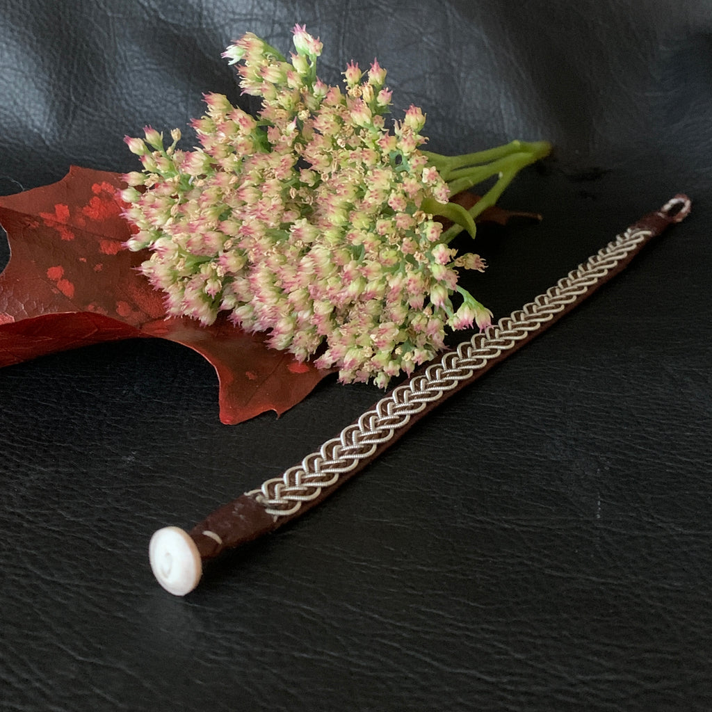 Tenntrådsarmband med flätning av tenntråd och tvinnat mörkbrunt garn på brunt renskinn. Knapp i renhorn och ögla i tvinnat renskinn. Bredd ca 10 mm. Samiskt hantverk av Doris Risfjell. Risfjells Sameslöjd.