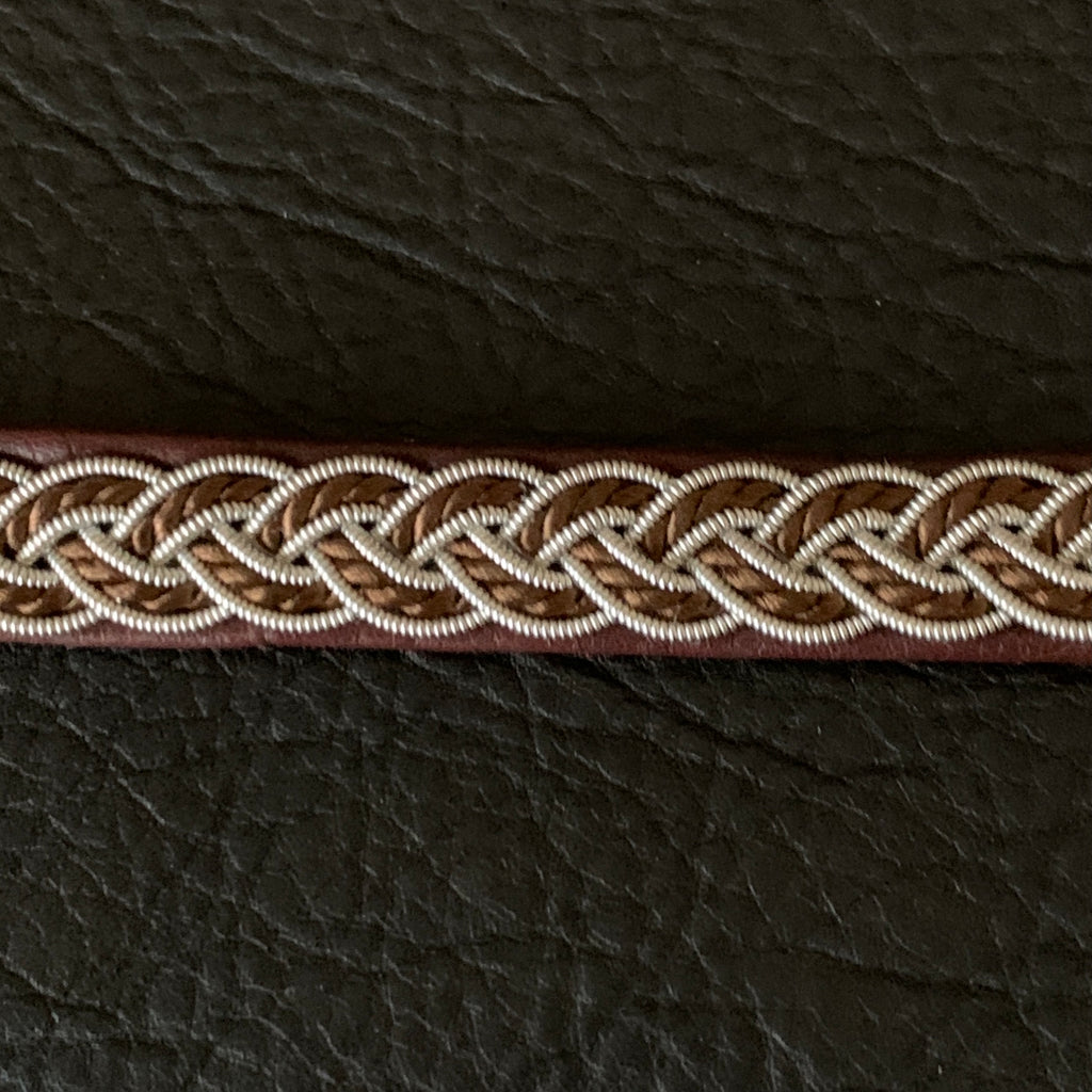 Tenntrådsarmband med flätning av tenntråd och tvinnat mörkbrunt garn på brunt renskinn. Knapp i renhorn och ögla i tvinnat renskinn. Bredd ca 10 mm. Samiskt hantverk av Doris Risfjell. Risfjells Sameslöjd.