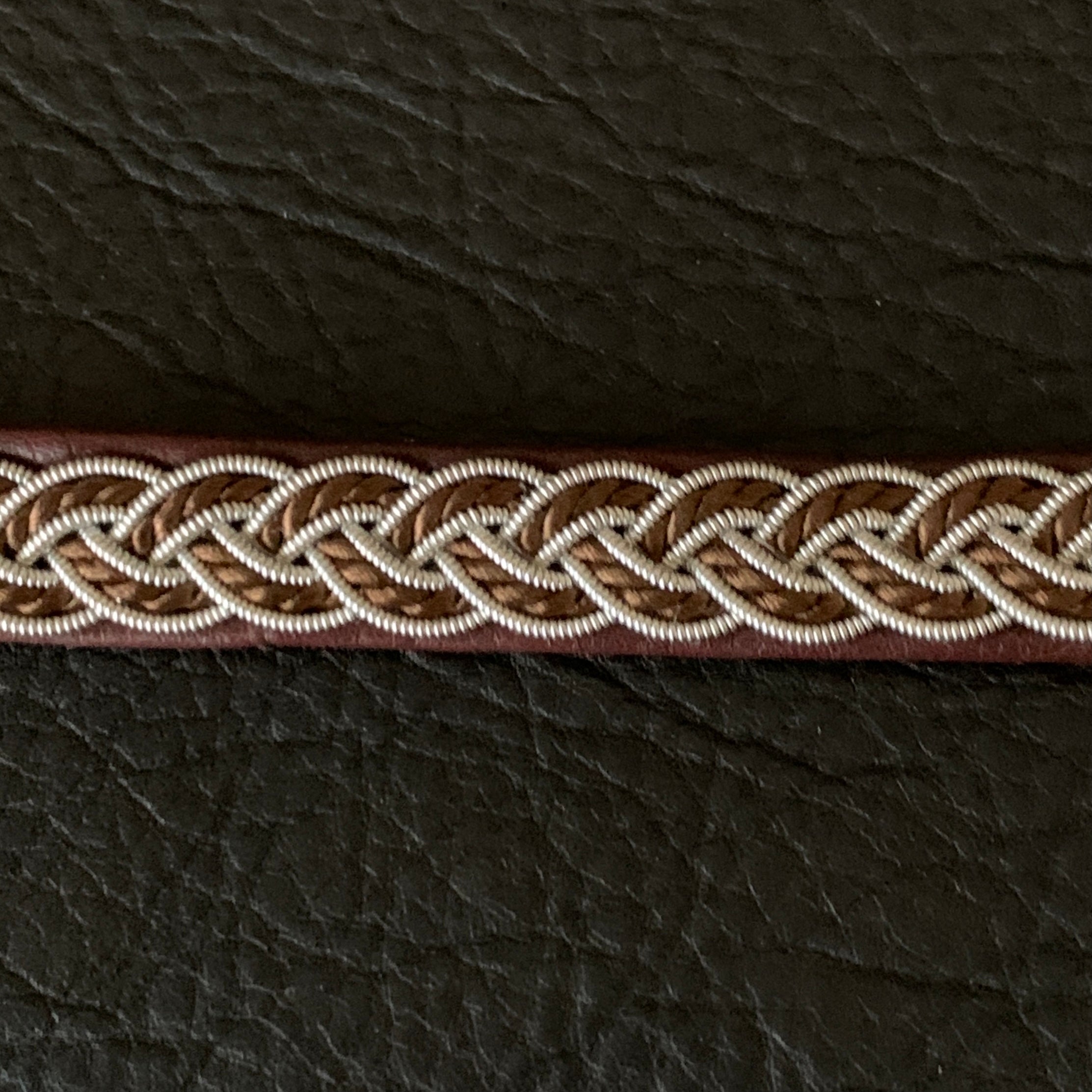 Tenntrådsarmband med flätning av tenntråd och tvinnat mörkbrunt garn på brunt renskinn. Knapp i renhorn och ögla i tvinnat renskinn. Bredd ca 10 mm. Samiskt hantverk av Doris Risfjell. Risfjells Sameslöjd.