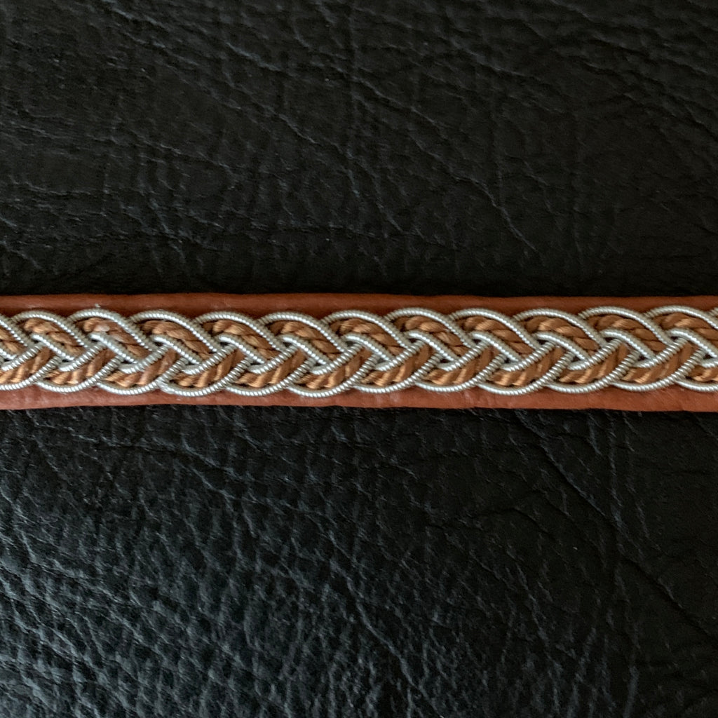 Tenntrådsarmband med flätning av tenntråd och tvinnat ljusbrunt garn på brunt renskinn med flätningar i tenntråd. Knapp i renhorn och ögla i tvinnat renskinn. Bredd ca 10 mm. Samiskt hantverk av Doris Risfjell.Risfjells Sameslöjd i Vilhelmina.