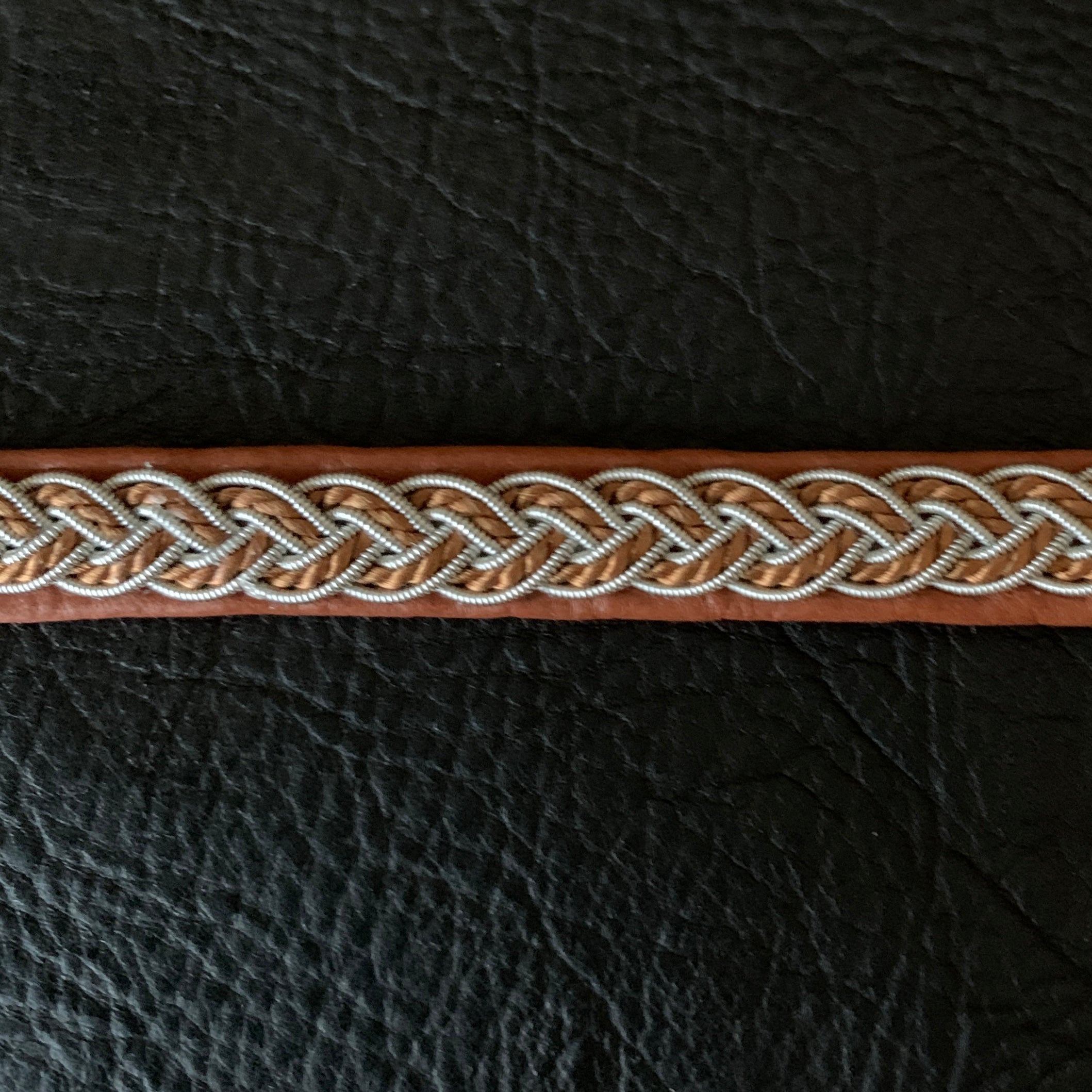 Tenntrådsarmband med flätning av tenntråd och tvinnat ljusbrunt garn på brunt renskinn med flätningar i tenntråd. Knapp i renhorn och ögla i tvinnat renskinn. Bredd ca 10 mm. Samiskt hantverk av Doris Risfjell.Risfjells Sameslöjd i Vilhelmina.