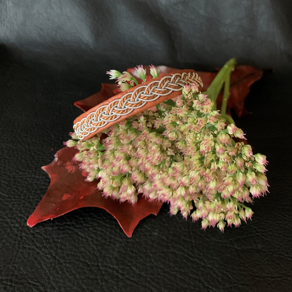 Tenntrådsarmband med flätning av tenntråd och tvinnat ljusbrunt garn på brunt renskinn med flätningar i tenntråd. Knapp i renhorn och ögla i tvinnat renskinn. Bredd ca 10 mm. Samiskt hantverk av Doris Risfjell.Risfjells Sameslöjd i Vilhelmina.