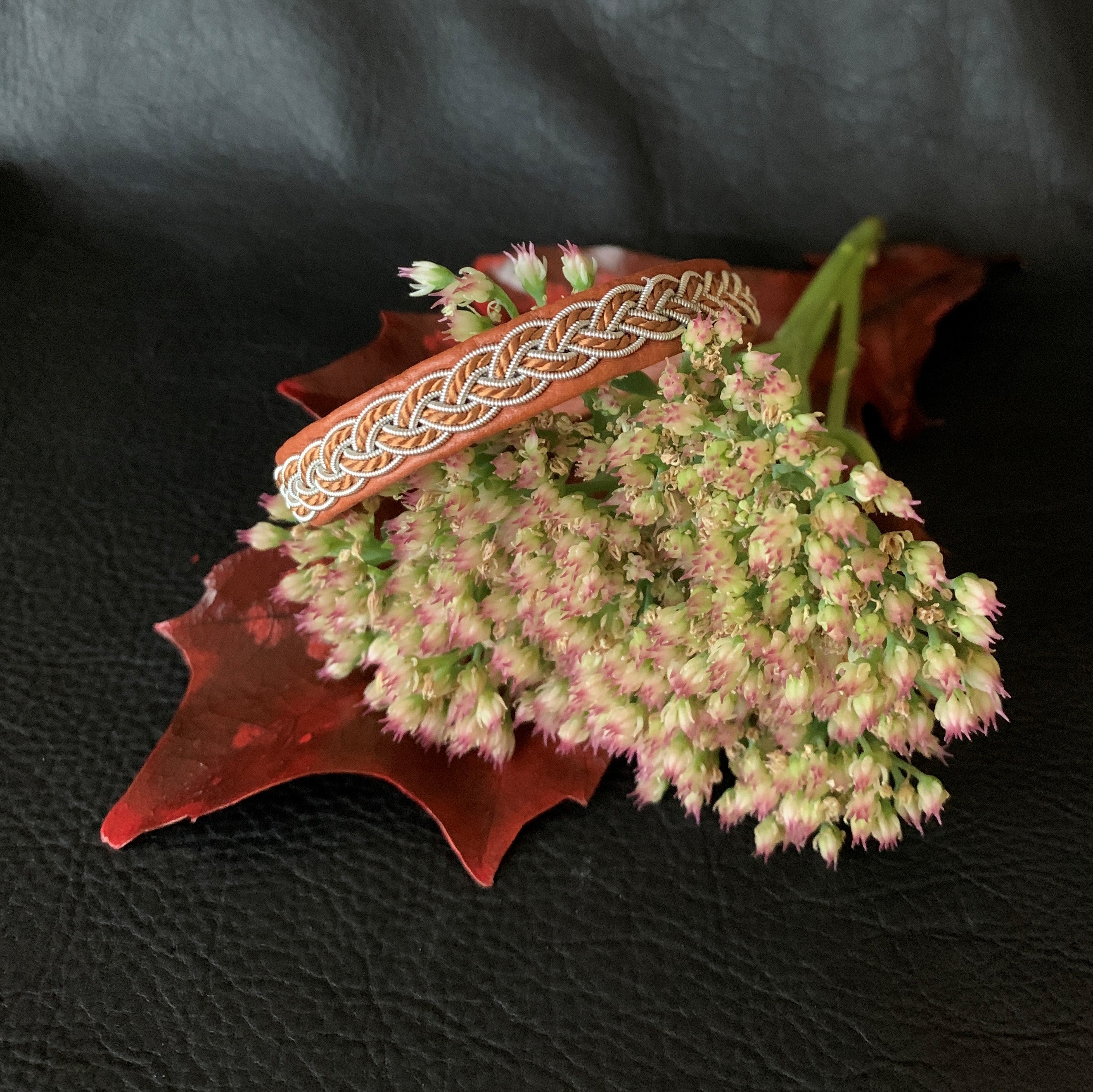 Tenntrådsarmband med flätning av tenntråd och tvinnat ljusbrunt garn på brunt renskinn med flätningar i tenntråd. Knapp i renhorn och ögla i tvinnat renskinn. Bredd ca 10 mm. Samiskt hantverk av Doris Risfjell.Risfjells Sameslöjd i Vilhelmina.