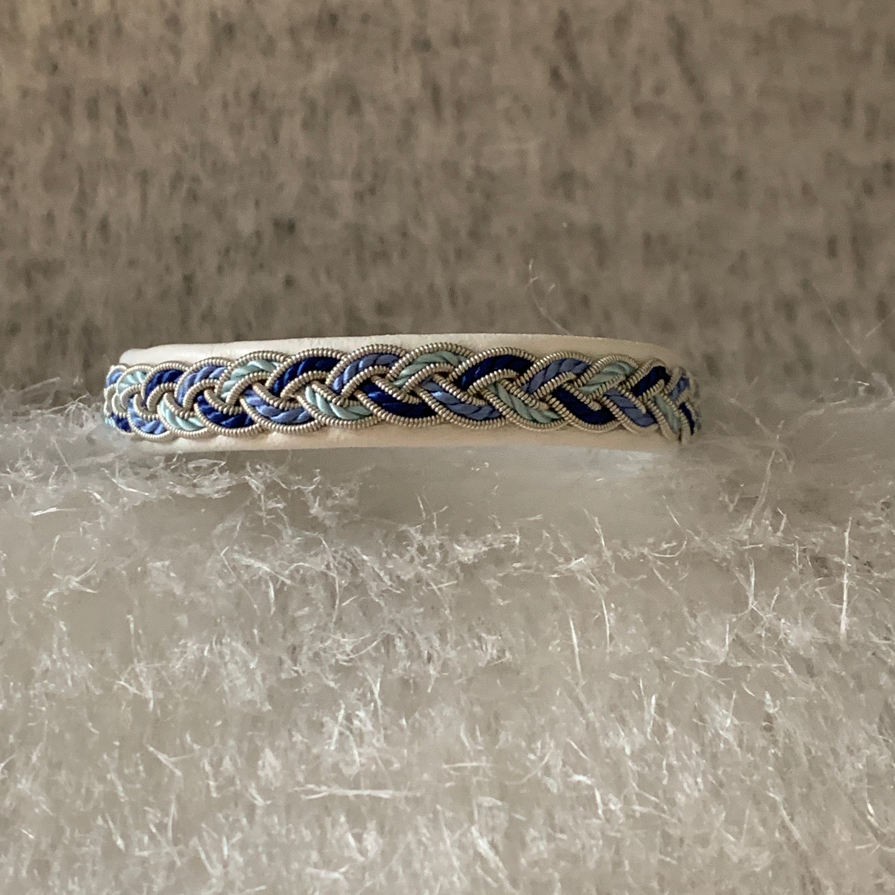 Tenntrådsarmband Vårjokk med flätning av tenntråd och blått, vitt och turkos tvinnat garn på vitt renskinn. Knapp i renhorn och ögla i tvinnat renskinn. Bredd ca 10 mm. Samiskt hantverk av Doris Risfjell, Risjfells Sameslöjd i Vilhelmina.
