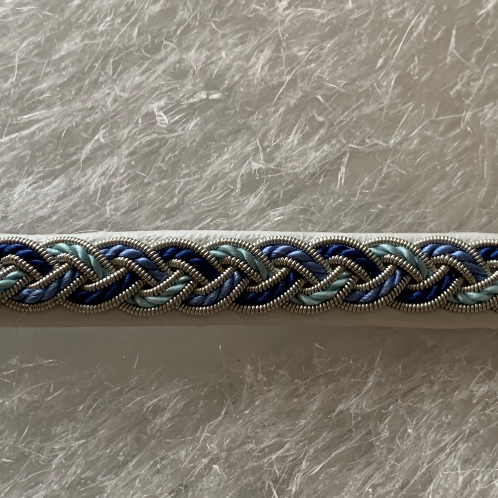 Tenntrådsarmband Vårjokk med flätning av tenntråd och blått, vitt och turkos tvinnat garn på vitt renskinn. Knapp i renhorn och ögla i tvinnat renskinn. Bredd ca 10 mm. Samiskt hantverk av Doris Risfjell.