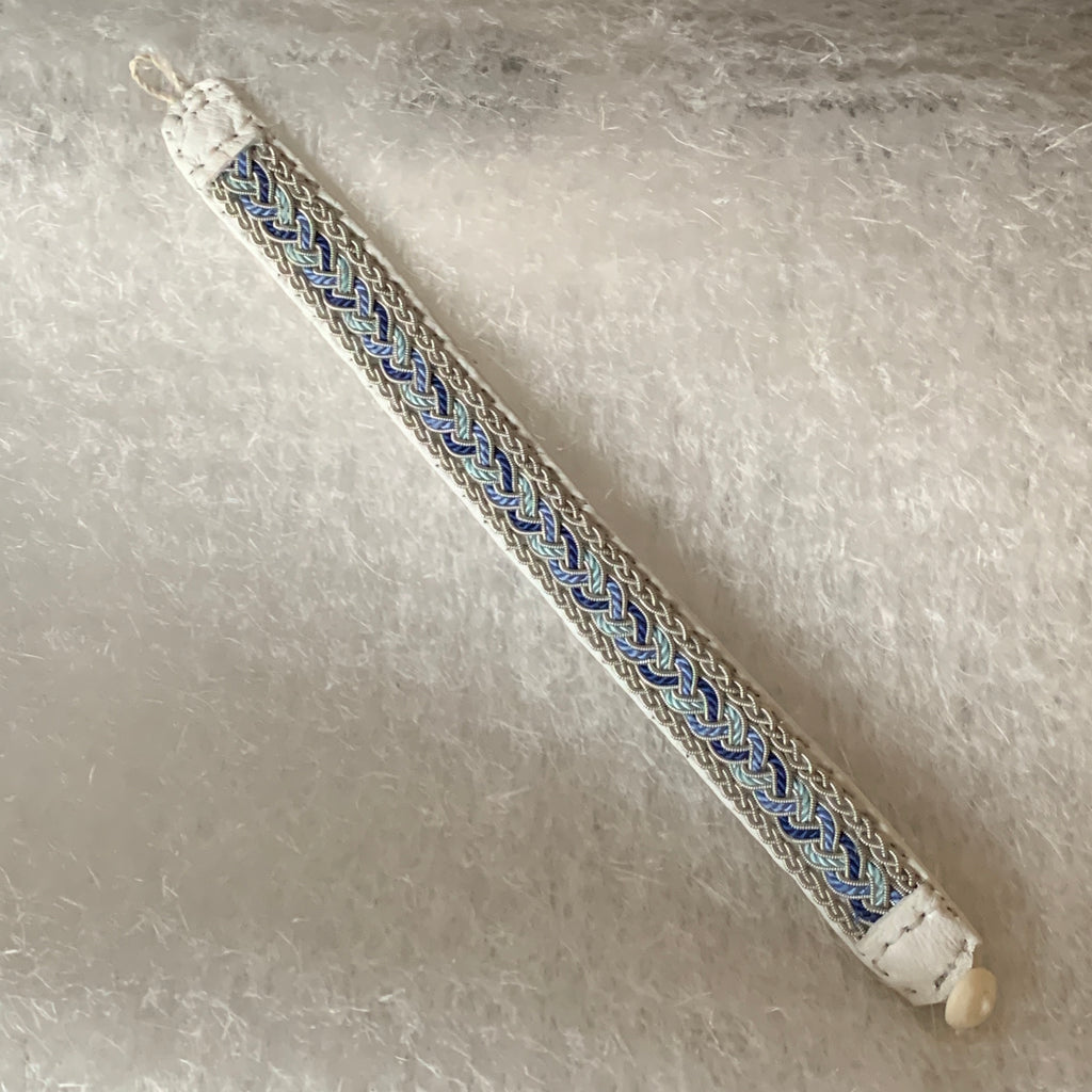 Tenntrådsarmband Vårjokk med flätning av tenntråd och blått, vitt och turkos tvinnat  på vitt renskinn. Knapp i renhorn och ögla i tvinnat renskinn. Bredd ca 10 mm. Samiskt hantverk av Doris Risfjell, Risjfells Sameslöjd i Vilhelmina.