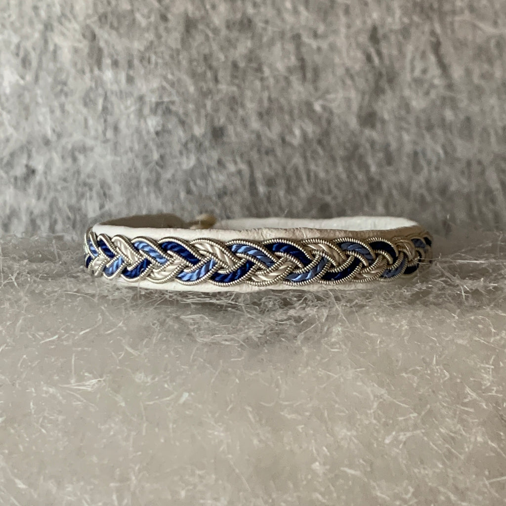 Tenntrådsarmband Vinterjokk med flätning av tenntråd och tvinnat blått och vitt garn på vitt renskinn. Knapp i renhorn och ögla i tvinnat renskinn. Bredd ca 10 mm. Samiskt hantverk av Doris Risfjell, Risfjells Sameslöjd Vilhelmina.
