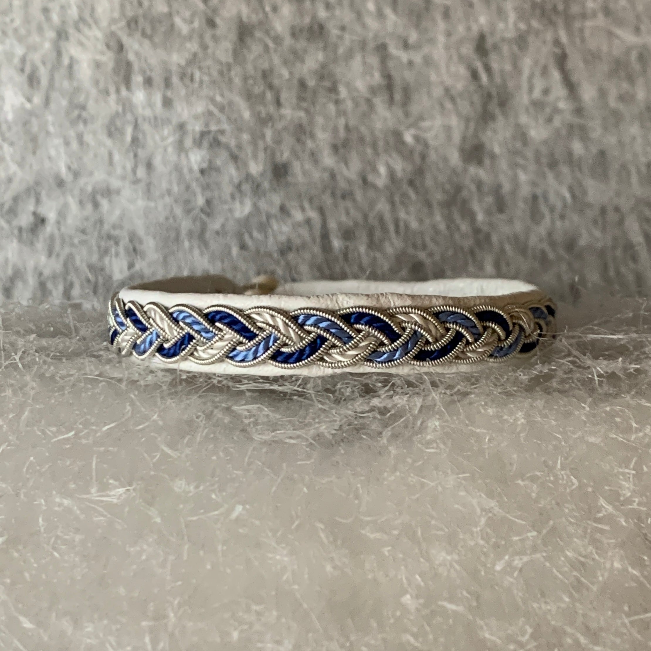 Tenntrådsarmband Vinterjokk med flätning av tenntråd och tvinnat blått och vitt garn på vitt renskinn. Knapp i renhorn och ögla i tvinnat renskinn. Bredd ca 10 mm. Samiskt hantverk av Doris Risfjell, Risfjells Sameslöjd Vilhelmina.