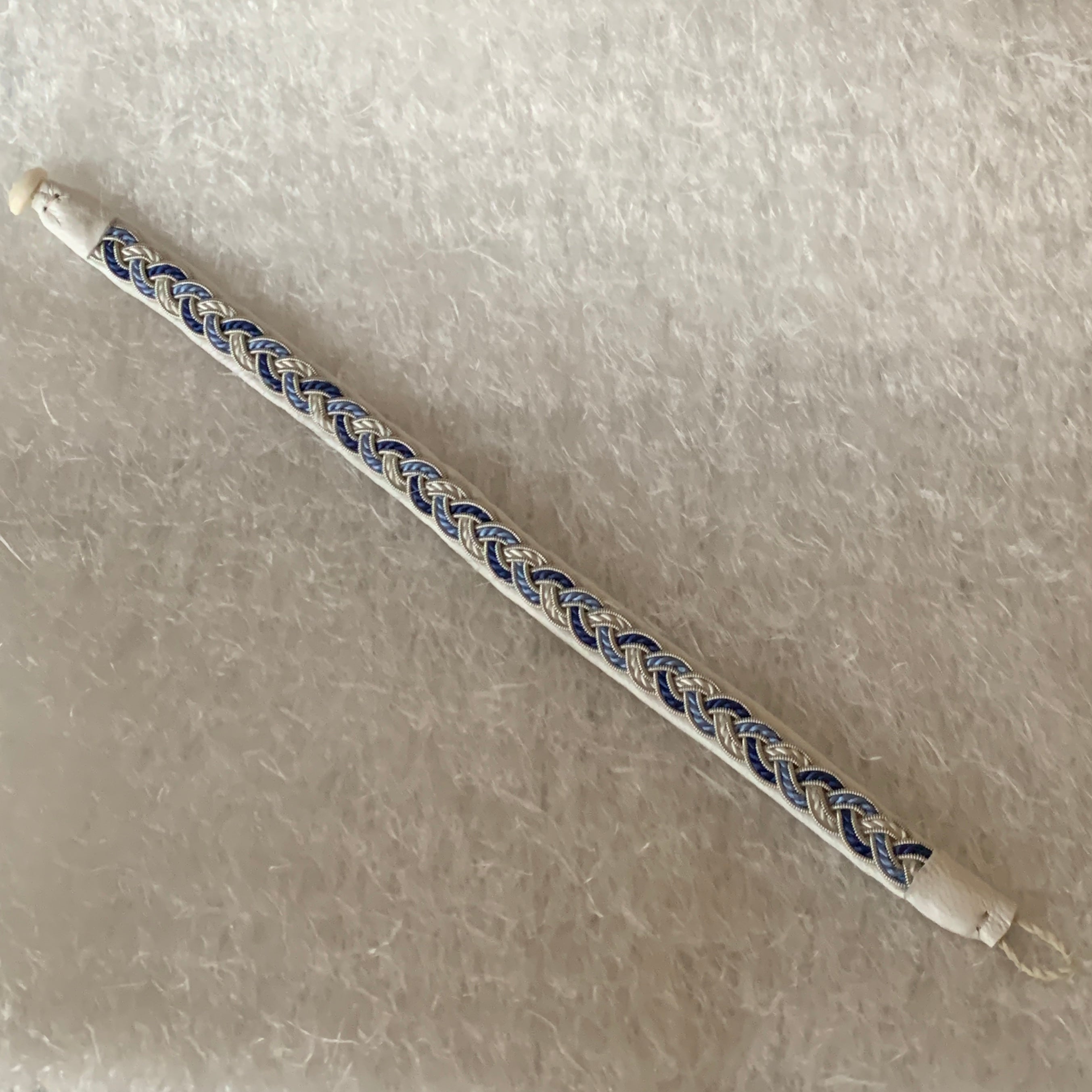 Tenntrådsarmband Vinterjokk med flätning av tenntråd och tvinnat blått och vitt garn på vitt renskinn. Knapp i renhorn och ögla i tvinnat renskinn. Bredd ca 10 mm. Samiskt hantverk av Doris Risfjell, Risfjells Sameslöjd Vilhelmina.